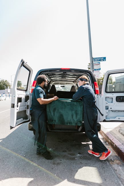 A man wearing a cap, glasses, and a dark vest over a blue jacket is engaged in a home relocation process, standing beside an open van with its rear doors wide open. Inside the van, there are multiple