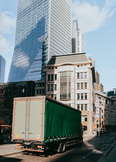 A large green cargo truck with closed side curtains is parked on a city street in front of tall modern office buildings with glass facades and a historic-style stone building with large windows. The truck is positioned near the curb, with its rear facing the viewer, indicating an active loading or unloading process typical of home relocation and furniture transport activities. The street scene includes pedestrians walking along the pavement and another vehicle in the distance, with clear weather and blue sky overhead. The setting reflects an urban environment suitable for professional removals, such as those offered by Man With a Van Brook Green, coordinating moving logistics in the Shepherds Bush area.