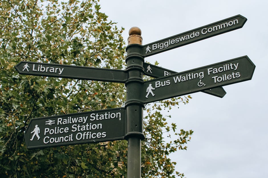 A black multi-directional street signpost set against a background of green foliage and an overcast sky. The signpost has six rectangular signs with white text and icons, pointing in different directi