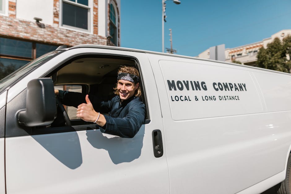 A man with glasses wearing a light blue shirt and dark trousers sits on a woven chair beside the open rear doors of a white van during a home relocation process. Inside the van, visible are large card