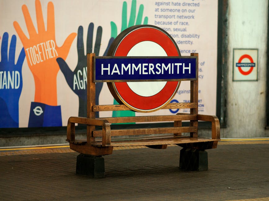 A wooden bench with wooden armrests and legs, positioned on a dark tiled platform inside Hammersmith underground station, featuring a circular London Underground sign with a red border and white cente