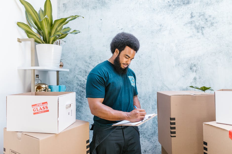 A man with dark, curly hair and a beard, dressed in a navy blue t-shirt, stands indoors surrounded by packed cardboard boxes of various sizes, some labeled with red 'Fragile' stickers, and writing on 